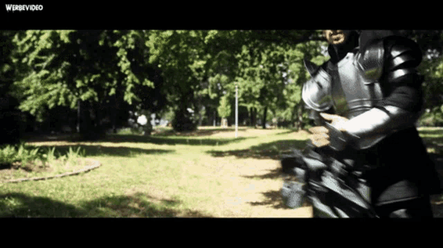 a man in armor is walking through a park with a webvideo watermark on the bottom right