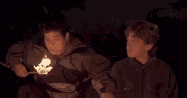 two young men are sitting around a fire in the dark .