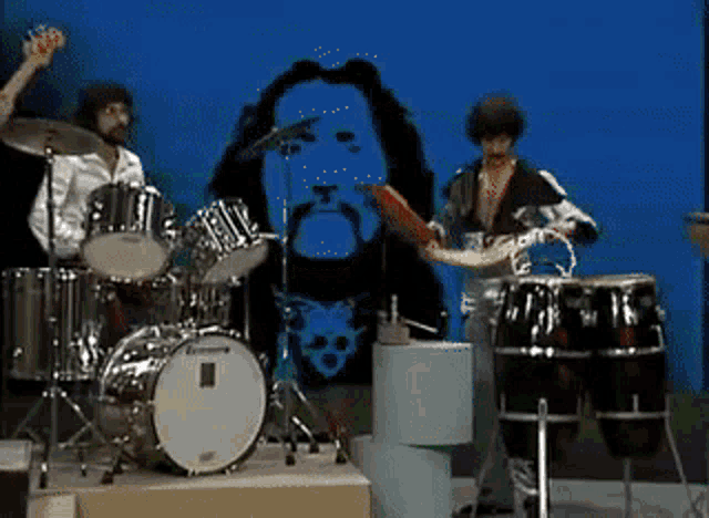 a man playing drums in front of a picture of a man with a beard