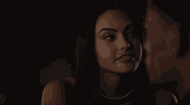 a woman wearing a pearl necklace looks at a man in a dark room