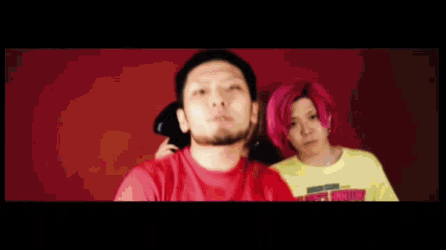 a man with a beard and a woman with pink hair are standing next to each other in front of a red wall .