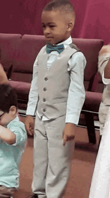 a young boy wearing a vest and bow tie stands in front of purple chairs