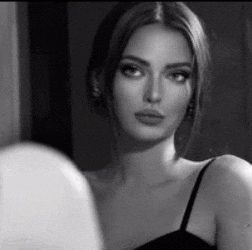 a black and white photo of a woman in a black dress looking at herself in the mirror .