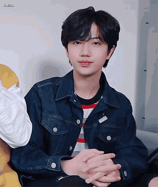a young man wearing a denim jacket is sitting with his hands folded in front of him