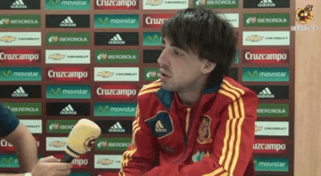 a man talking into a microphone in front of a wall with advertisements for cruzcampo