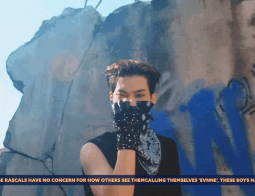 a man wearing a bandana and gloves is standing in front of a wall with graffiti