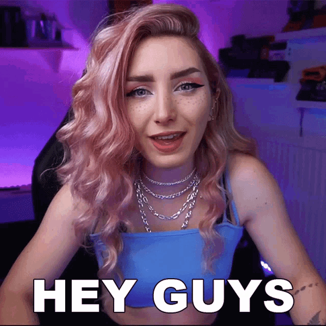 a woman with pink hair says " hey guys " while wearing a blue top