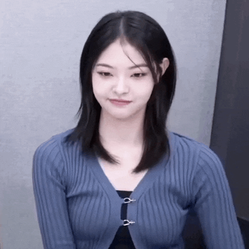 a young woman wearing a blue sweater is sitting in front of a wall and looking at the camera .