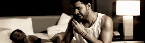 a man in a white tank top is sitting on a bed talking on a cell phone