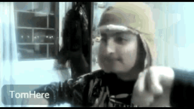 a person wearing a hat and a black shirt is pointing at the camera with the words tomhere written below them