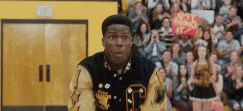 a man in a letterman jacket is standing in front of a crowd of people in a gym .