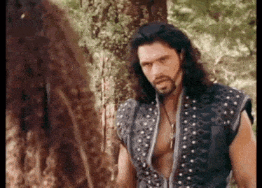 a man with long hair and a beard is standing next to a tree in the woods .