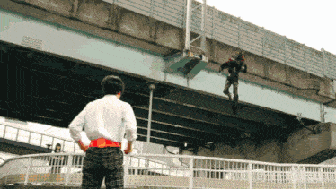 a man in a white shirt and plaid pants stands on a bridge watching a person hanging from a bridge