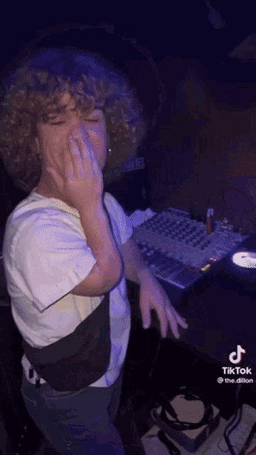a little boy with curly hair is standing in front of a mixing board and covering his face with his hand .