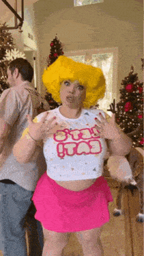 a woman in a yellow wig and pink skirt is standing in front of a christmas tree in a room .