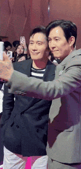 two men in suits are taking a selfie together in front of a crowd of people .