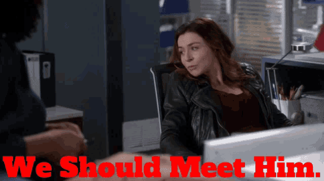 a woman in a leather jacket sits at a desk with the words we should meet him