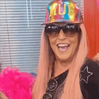a woman wearing a hat , sunglasses and a pink wig is holding a pink pom pom .