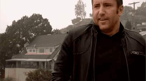 a man in a black leather jacket is standing in front of a house on a rainy day .