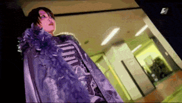 a woman in a purple fur coat is standing in a hallway with the letter k on the bottom right