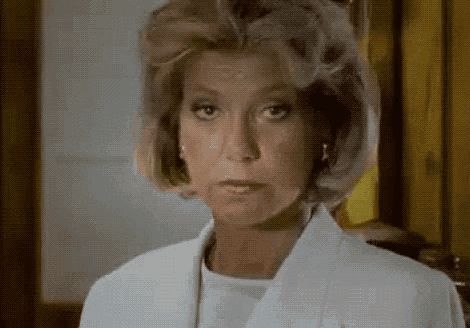 a woman wearing a white jacket and earrings is looking at the camera with a serious look on her face .