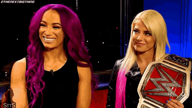 two women with purple hair and blonde hair are standing next to each other holding a wrestling championship belt .