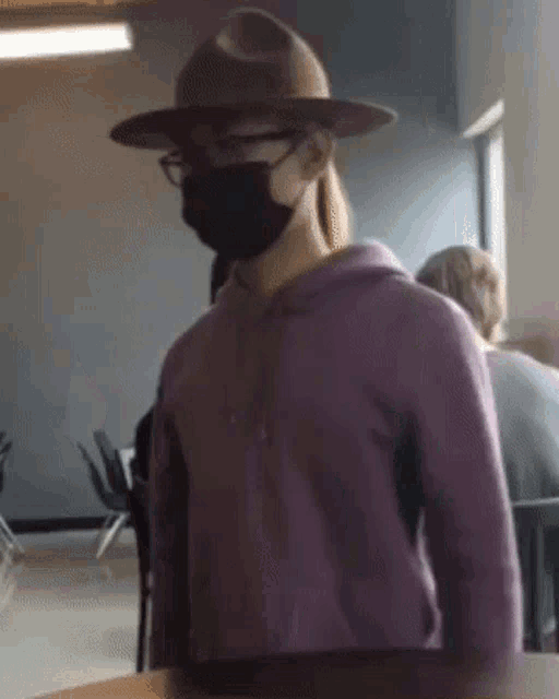 a person wearing a hat and a mask is sitting at a table in a room .