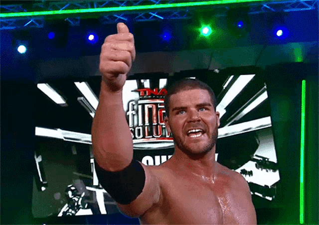 a shirtless wrestler giving a thumbs up in front of a tna logo