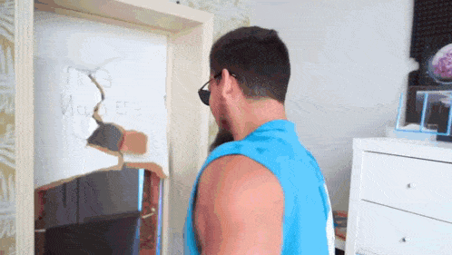 a man in a blue tank top is standing in front of a door that has a hole in it that says " no "