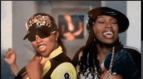 two women are standing next to each other in a room . one of the women is wearing sunglasses and a hat .
