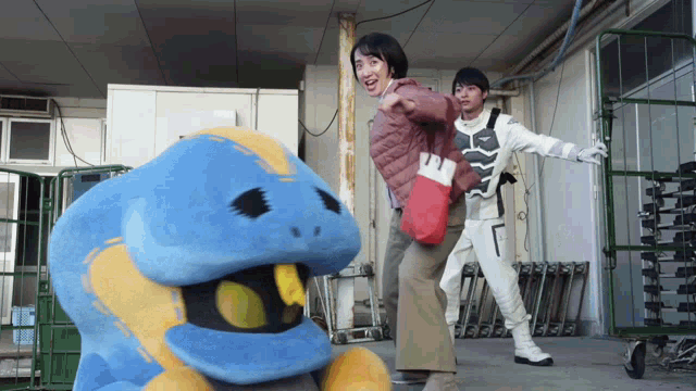 a woman in a pink jacket is standing next to a blue and yellow stuffed animal
