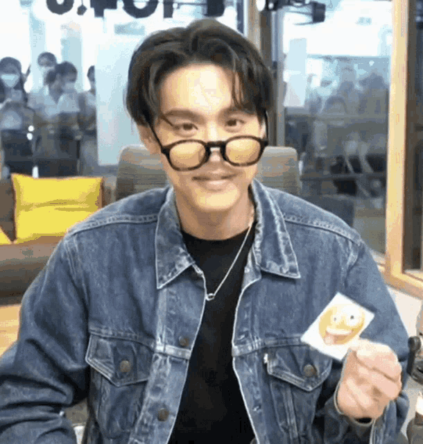 a man wearing glasses and a denim jacket is holding a smiley face sticker .
