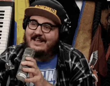 a man with a beard and glasses is holding a microphone and wearing a hat with the word killer on it