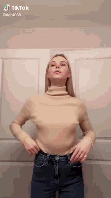 a woman is standing in front of a door wearing a tan turtleneck and jeans .