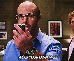 a bald man wearing glasses is holding a cell phone to his mouth and says " fuck your own face "