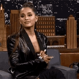 ariana grande is sitting on a couch wearing a black jacket and holding a cell phone .