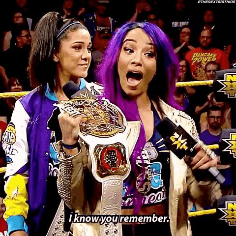 a woman with purple hair is holding a championship belt and talking into a microphone while another woman looks on .