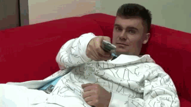 a man in a white jacket is laying on a red couch holding a remote control .