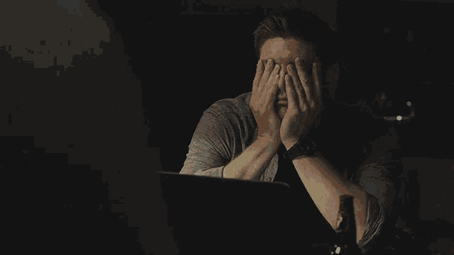 a man is sitting in front of a laptop computer covering his face with his hands .