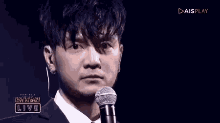 a man in a suit stands in front of a microphone with aisplay on the bottom right