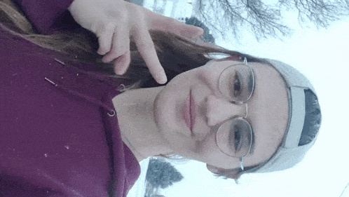 a woman wearing glasses , a hat and a purple hoodie is standing in the snow .
