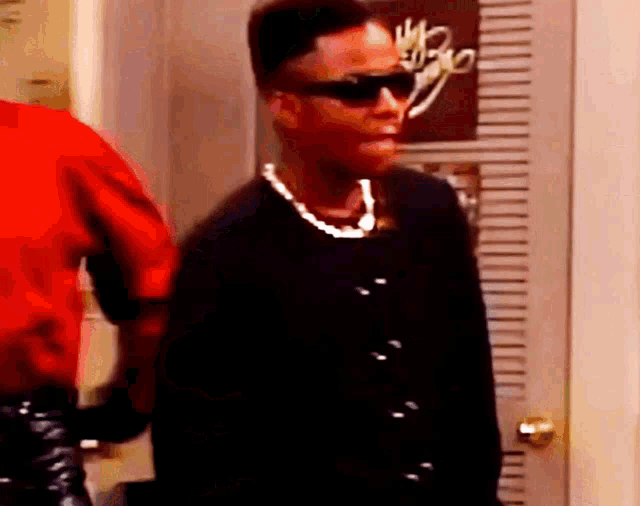 a man wearing sunglasses and a pearl necklace is standing in a room .