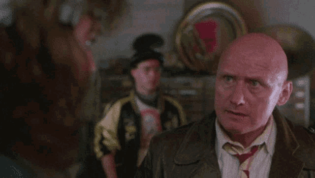 a bald man in a suit and tie is talking to a woman while another man looks on .