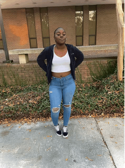 a woman wearing ripped jeans and a white crop top stands on a sidewalk