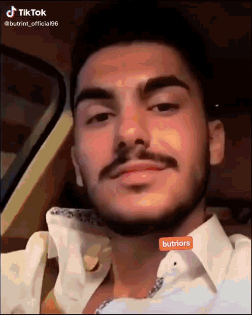 a man with a beard is sitting in a car and has a tiktok sticker on his neck that says buttriors
