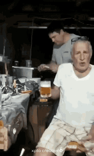a man is holding a glass of beer while another man prepares food in the background