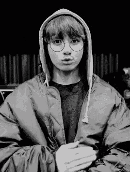 a black and white photo of a young man wearing a hooded jacket and glasses .