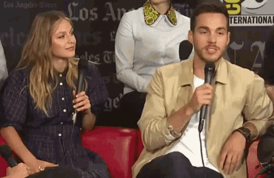 a man is holding a microphone while sitting next to a woman in front of a los angeles times sign