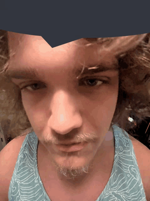 a man with curly hair and a beard wearing a blue tank top looks at the camera