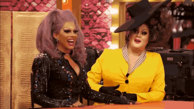two drag queens are sitting at a table and one is wearing a yellow suit